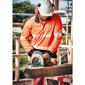 A worker in an orange segmented tape long sleeve polo shirt operates a circular saw at a construction site.