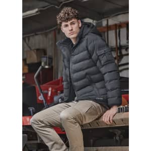 A unisex hooded puffer jacket in black, featuring a fitted design and a logo, worn by a model sitting on machinery.