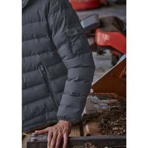 A grey unisex puffer jacket with a zip and pocket detail on the sleeve, displayed on a person near machinery.
