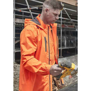 A man wearing an orange unisex packable rain jacket, focused on using a power tool on a construction site.