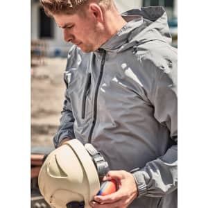 A man in a light grey rain jacket holds a safety helmet and looks down. The jacket features a logo.