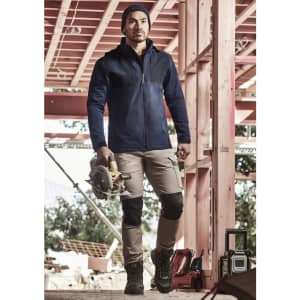 A man wearing a navy waterproof jacket stands on a construction site, holding a saw. He pairs it with beige pants and boots.