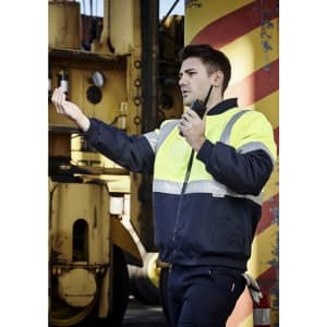 A man in a hi-vis quilted flying jacket, featuring yellow and navy colours, standing with a radio in a work environment.