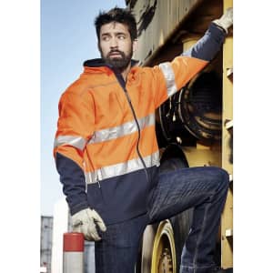 A unisex hi-vis softshell jacket in orange and navy, featuring reflective strips, worn by a person leaning against machinery.