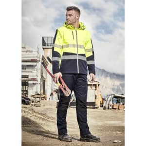 A man in a high-visibility yellow softshell jacket stands on a construction site, holding tools. The jacket features reflective strips.