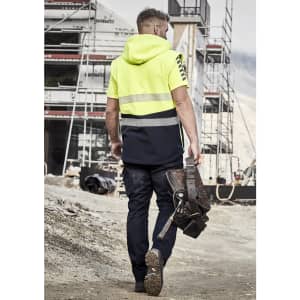 A man in a high-visibility yellow and navy softshell jacket walks at a construction site, holding a tool belt.