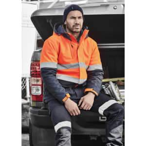 A man wearing an orange and navy unisex hi-vis softshell jacket sits on the back of a vehicle.