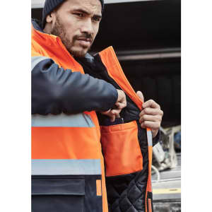A man in an orange and navy softshell jacket with reflective strips, reaching into an inner pocket.