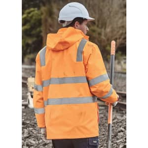 A person wearing an orange waterproof jacket with reflective stripes and a white hard hat, standing near railway tracks.