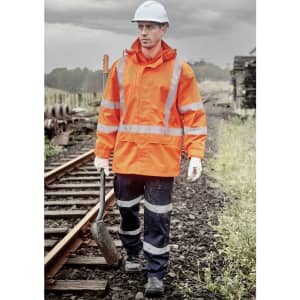 A man wearing an orange waterproof jacket and navy trousers walks along railway tracks, holding a tool. The jacket features reflective stripes.