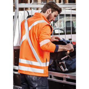 An orange unisex softshell jacket with reflective strips worn by a person at a worksite, with tools nearby.