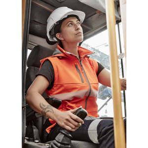 A woman wearing an orange softshell jacket with reflective strips, seated in a construction vehicle, wearing a hard hat.