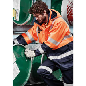 A man in an orange flame-resistant jacket adjusts a green barrel, wearing gloves and dark trousers.