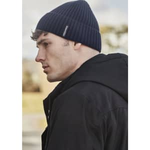 A side view of a man wearing a navy ribbed beanie, paired with a black jacket, outdoors. The beanie features a logo.