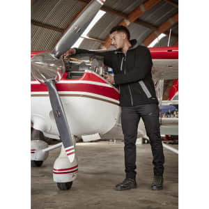 A man in black cargo pants works on an aircraft engine inside a hangar, wearing a black fleece jacket.