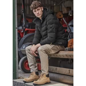 A man sits on a bench wearing khaki cargo pants and a black puffer jacket, with tan boots, in a rustic setting.