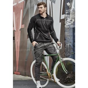 A man in black pullover and grey cuffed stretch pants poses with a green bicycle against a graffiti wall.