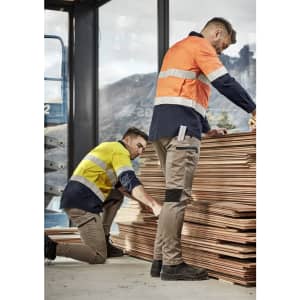 Two men in high-visibility work shirts and stretch pants, one crouching beside stacked timber, the other inspecting the wood.