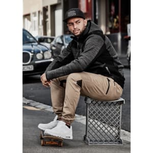 A man sits on a crate wearing cuffed beige stretch pants and a black hoodie. He has white sneakers and a cap.