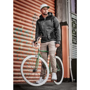 A man wearing cuffed beige stretch pants and a grey jacket stands beside a green bicycle against a textured orange backdrop.