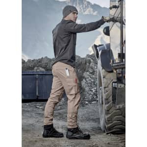Men's curved cargo pants in beige, featuring multiple pockets, paired with dark boots and a grey beanie against a rugged backdrop.