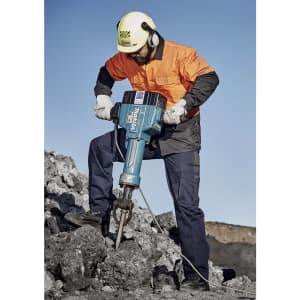 A man in safety gear operates a jackhammer on a rocky surface, wearing an orange shirt and navy blue cargo pants.