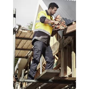 A man wearing a high-visibility yellow and black shirt and navy cargo pants uses a tool while standing on scaffolding.