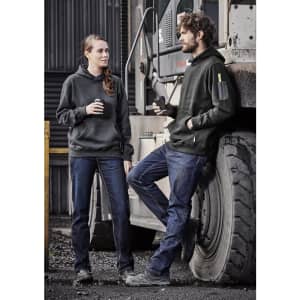 A man and woman wear black hoodies and blue denim work jeans, standing beside a large vehicle in an industrial setting.