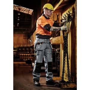 A man in an orange and grey shirt and multi-pocket grey pants works in a warehouse, wearing safety gear and a helmet.