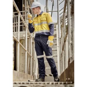 A worker in a yellow and navy work shirt and cargo pants, wearing gloves and a hard hat, on a metal staircase.