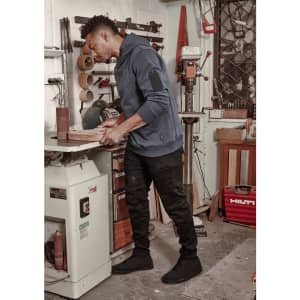 A man wearing a navy blue hoodie and black stretch jeans stands in a workshop, handling a piece of wood.