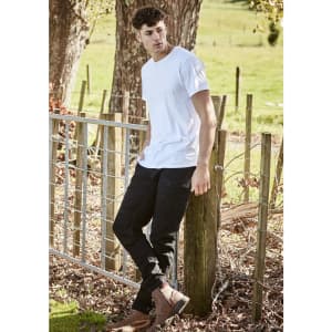 A person wearing black stretch jeans and a white t-shirt stands by a wooden fence in a grassy area.