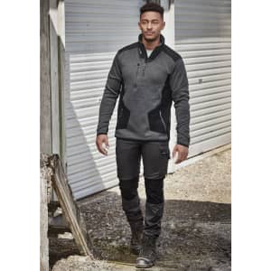 A man in dark grey and black workwear pants and a fitted grey pullover, walking in an outdoor setting.