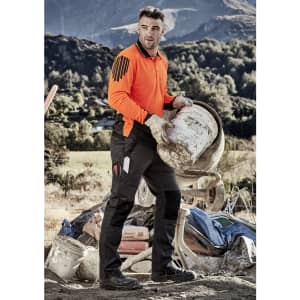 A man in an orange shirt and black tough work pants carries a bucket at a construction site, with mountains in the background.