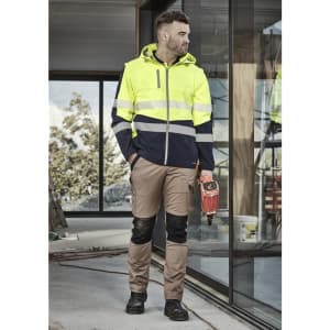 A man wearing a high-visibility yellow and navy work jacket and khaki work pants, standing in a construction setting.