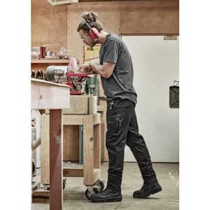 A person wearing black rugged cooling stretch pants stands in a workshop, using a tool. The pants feature pockets and have a logo.
