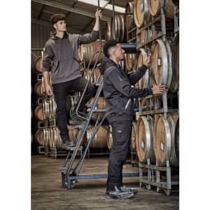 Two men in rugged cooling stretch pants, one standing on a ladder and the other inspecting wooden barrels, in a warehouse setting.