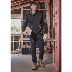 A man wearing black cargo pants and a black hoodie is holding wooden blocks, standing in a construction setting.