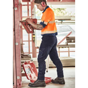 A worker in high-visibility orange shirt and navy cargo pants uses a tool on a construction site. Features branding.