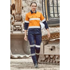 A woman wearing orange and navy cargo pants with reflective stripes, standing near heavy machinery. A hard hat is on her head.