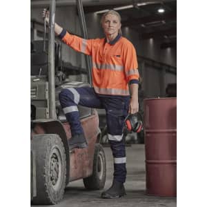 A woman in an orange high-visibility shirt and dark cargo pants leans against a forklift, wearing safety gear.