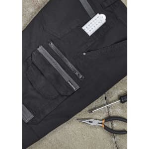 A pair of black work pants featuring multiple pockets, a zipper, and a branding logo. Tools are placed beside them.