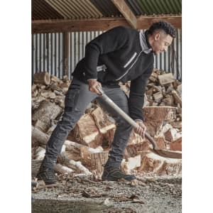 A model is wearing black work pants and a black fleece top while chopping wood in a rustic setting.