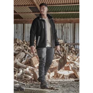 A model wearing black work pants and a black fleece jacket stands outdoors, with stacks of timber in the background.