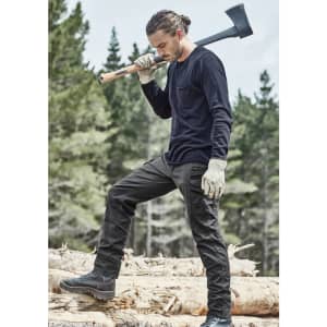 A man in black work pants and a long-sleeve top, holding an axe while standing on logs in a forested area.