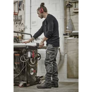 A worker in black Mens Streetworx Heritage Pants, operating machinery in a workshop, with tools and equipment in the background.