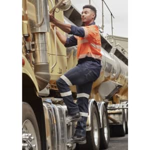 A man in an orange and navy work shirt climbs onto a truck, wearing dark blue stretch jeans with reflective stripes.