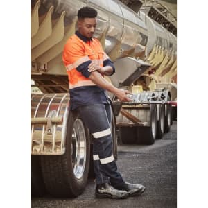 A man in a high-visibility orange and navy work shirt and dark stretch jeans leans against a truck.