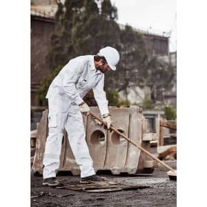 A man in white work pants and a helmet uses a broom at a construction site. The pants have a logo and pockets.