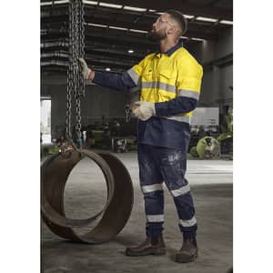 A man in a high visibility yellow and navy cargo pant with cuffs stands in an industrial setting, holding chains.
